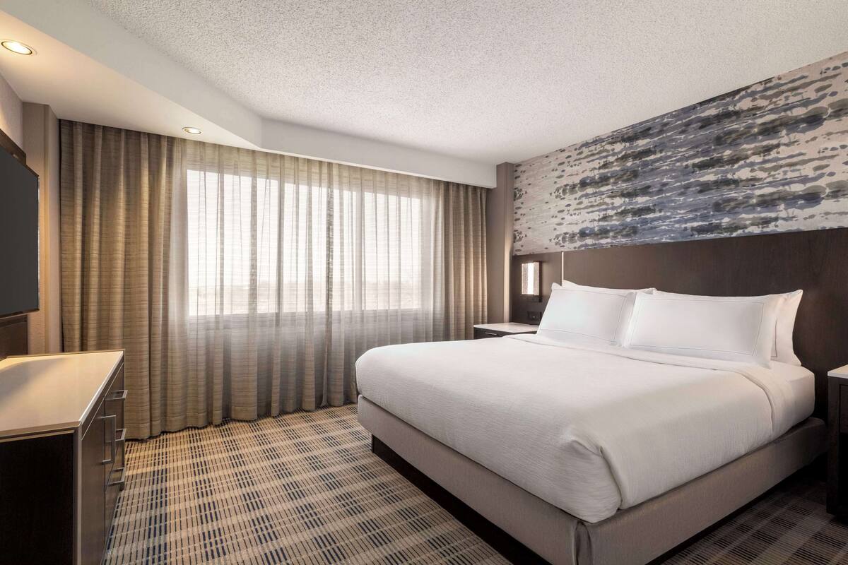 Ein Hotelzimmer mit einem großen Bett, einem Schreibtisch und einem großen Fenster mit Vorhängen.