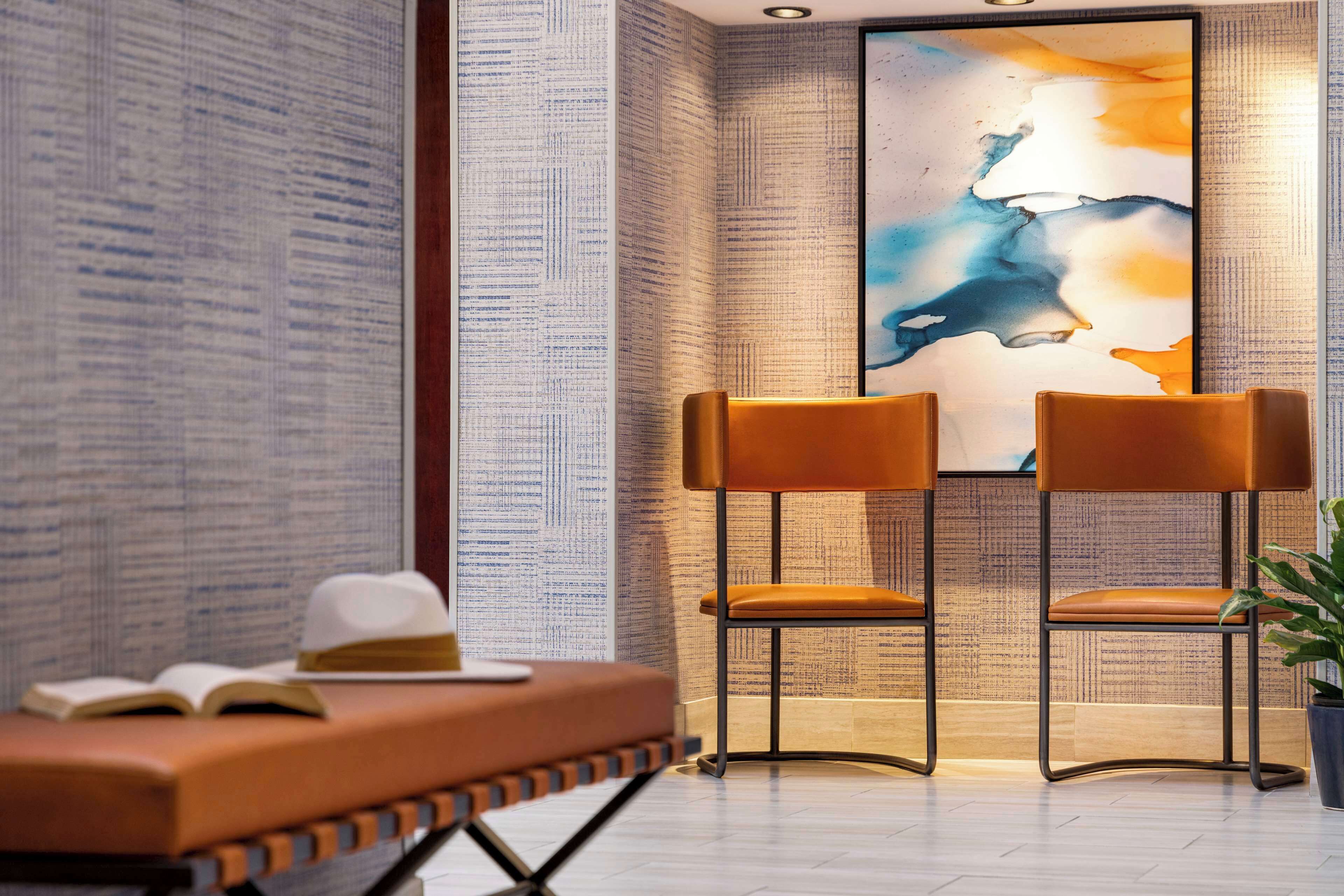 A modern room with a large abstract painting, two brown chairs, a small table with a book and a hat, and a potted plant.