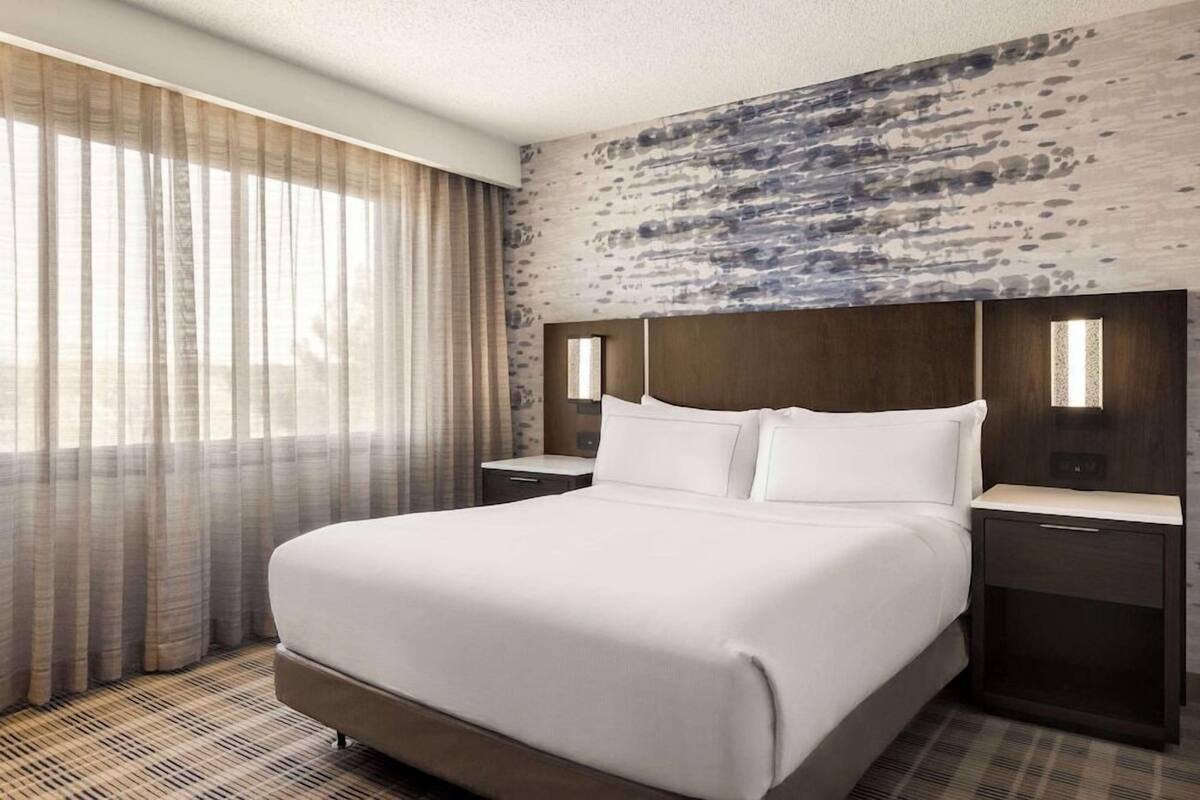 Ein Hotelzimmer mit einem großen Bett, zwei Nachttischen, einem Bettgestell mit strukturierter Oberfläche und durchsichtigen Vorhängen am Fenster.