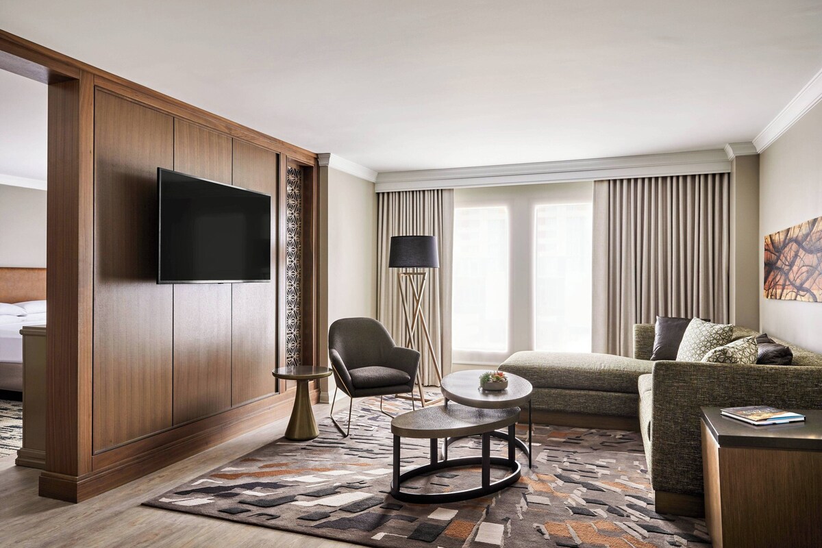 Habitación de hotel con armario de madera, televisión de pantalla plana, sofá, mesa de centro redonda, silla, lámpara de pie y alfombra estampada.
