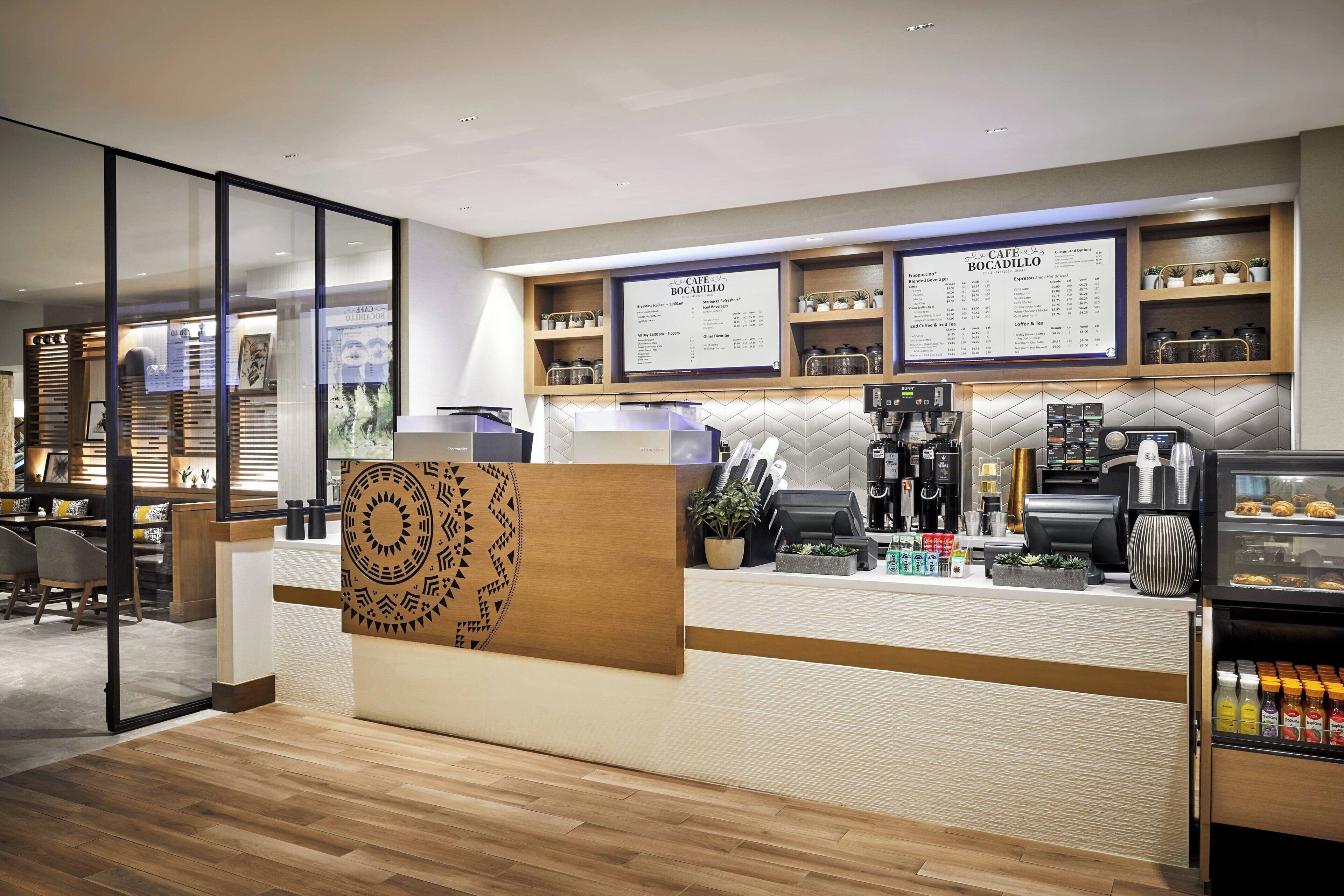 A modern cafe with a wooden reception desk, menu boards, coffee machines, and a display case with pastries.