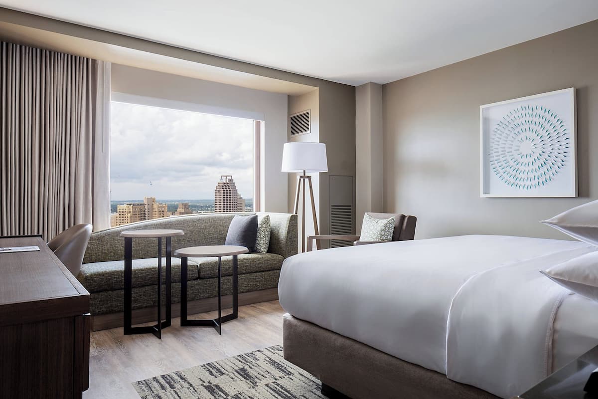 Une chambre d’hôtel avec un grand lit, un canapé, une petite table et une lampe. Il y a une fenêtre donnant sur des immeubles.