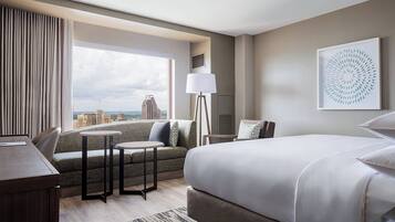 A hotel room with a large bed, a sofa, a small table, a lamp, and a city view.