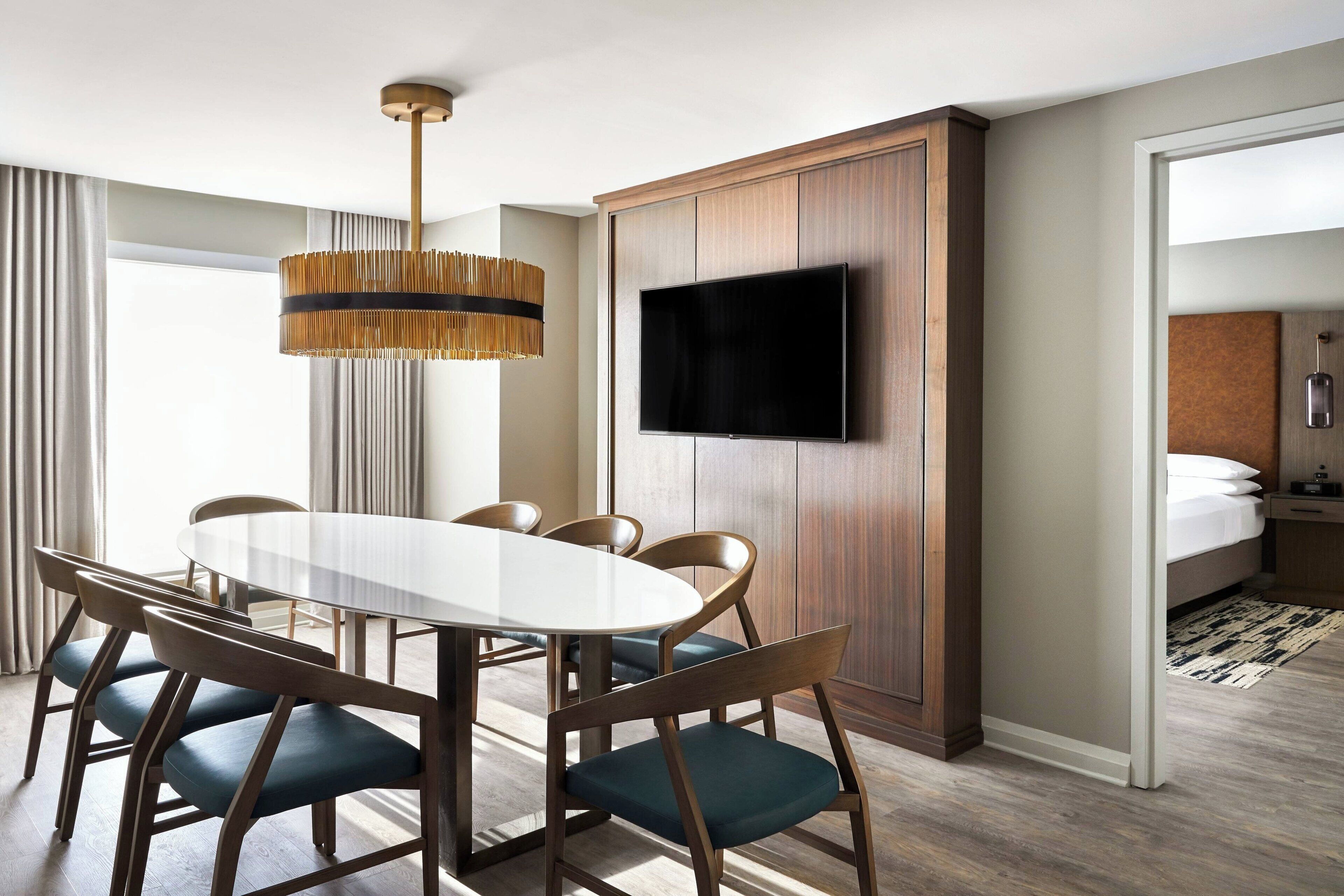 A hotel room with a dining area featuring a round table, chairs, a television mounted on a wooden panel, and a bed with a nightstand.