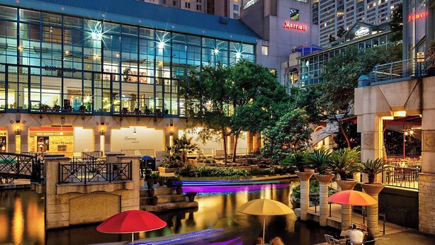 Un hotel con fachada de vidrio moderna, zona de estar exterior con sombrillas y un elemento decorativo de agua.