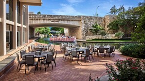 Un espacio de comedor al aire libre con mesas y sillas, un puente de piedra y un canal al fondo.