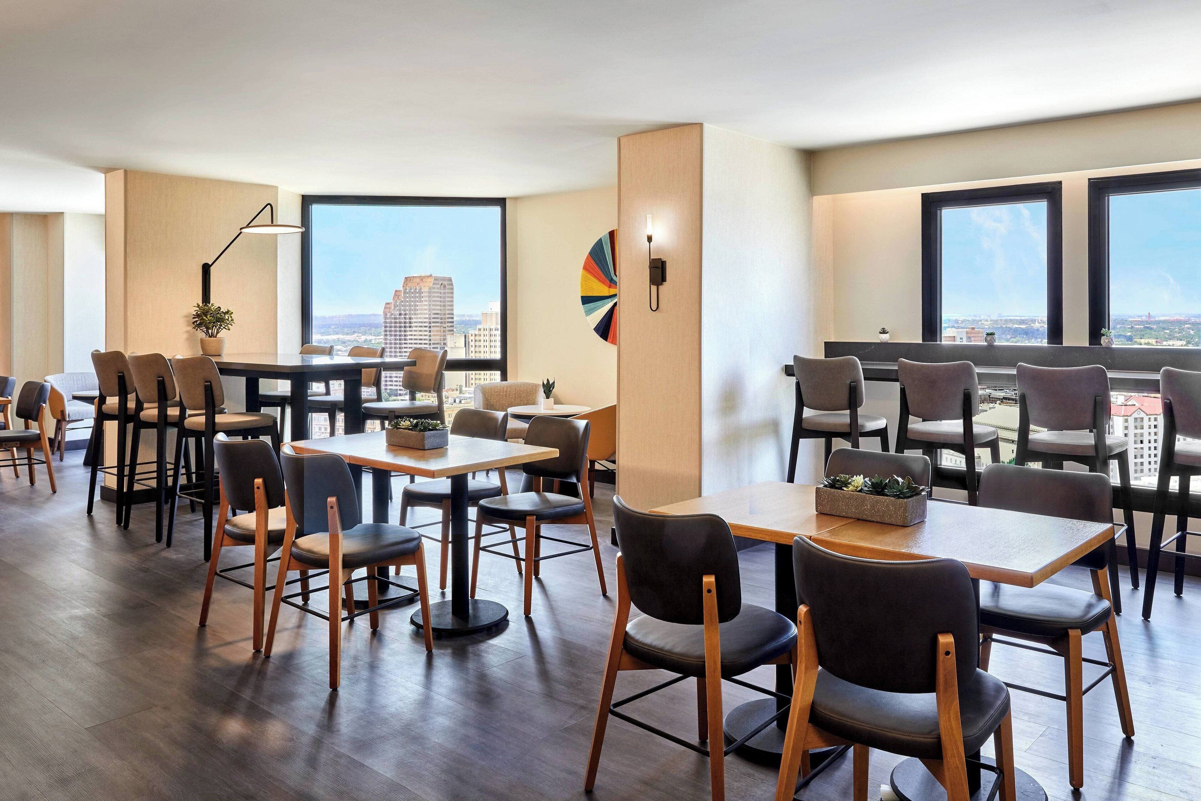 Un espace de restauration moderne, doté de plusieurs tables et chaises, de grandes fenêtres offrant une vue sur la ville et d’un élément décoratif mural.