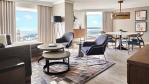 Habitación de hotel moderna con sofá, dos sillones, una mesa de comedor y un ventanal con vistas a la ciudad.