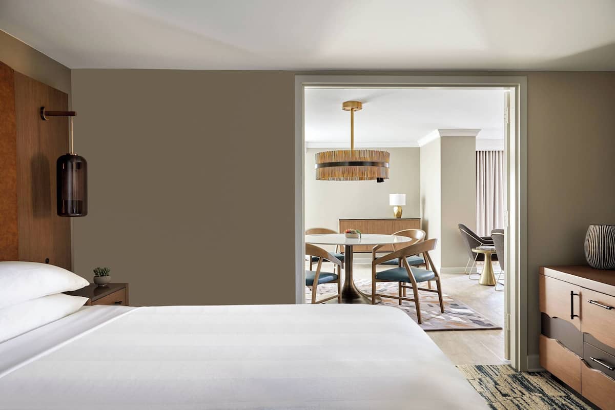 Une chambre d’hôtel avec un grand lit, une table de chevet en bois, une petite table avec une plante et un coin repas agrémenté d’un lustre.