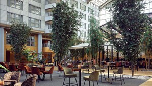 A modern indoor dining area with multiple tables and chairs, surrounded by large indoor plants and a high ceiling with natural light. - Van der Valk Hotel Paris CDG Airport (Roissy-en-France)