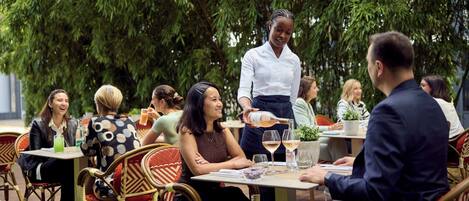 Un grupo de personas sentadas en una mesa al aire libre con sillas de mimbre, conversando animadamente. Un camarero sirve bebidas.