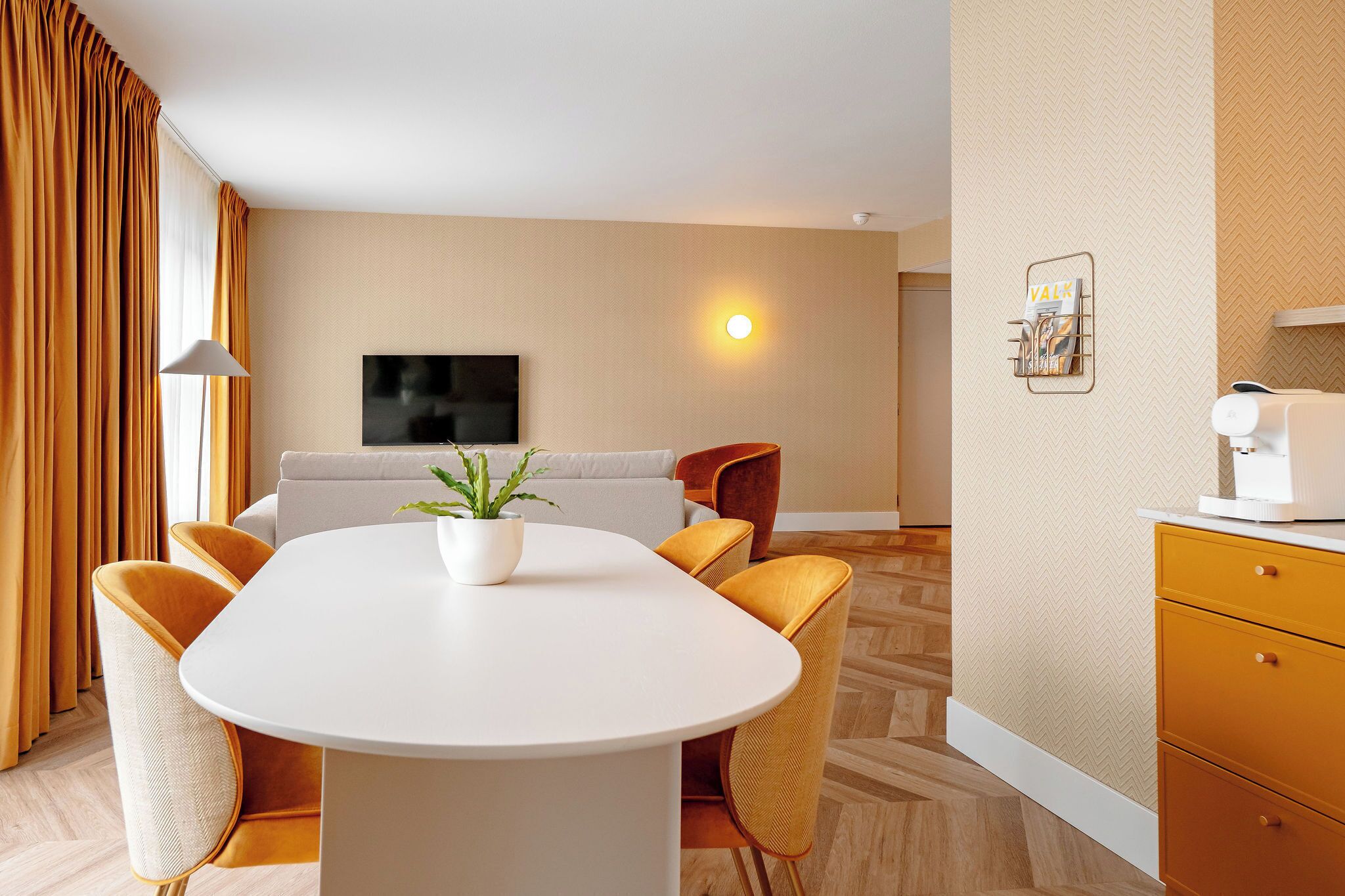 A modern hotel room with a round dining table, comfortable seating, a flat-screen TV, and a wall-mounted light fixture.