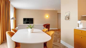 A hotel room with a dining table, chairs, a sofa, a TV, a wall-mounted coffee machine, and a potted plant.