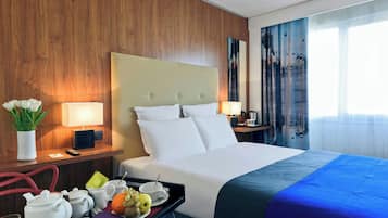 Habitación de hotel con una cama grande, paredes de paneles de madera, una mesita de noche con lámpara, una bandeja con alimentos para el desayuno y una ventana con cortinas.
