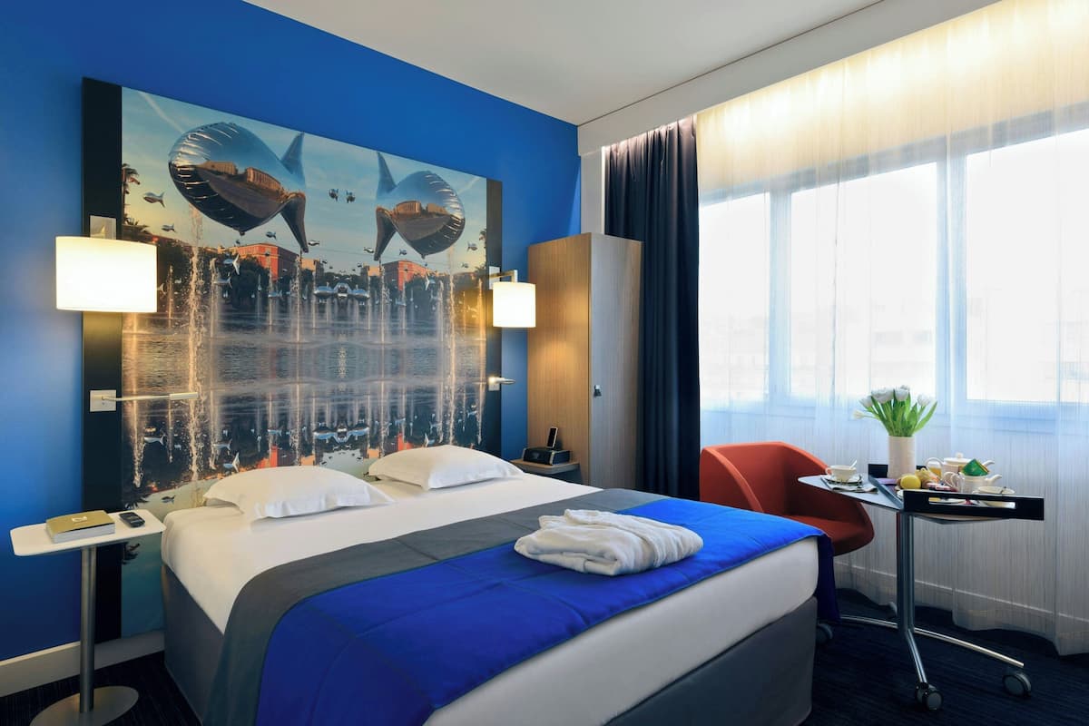 A hotel room with a large bed, a desk with a lamp, a chair, and a window with curtains.