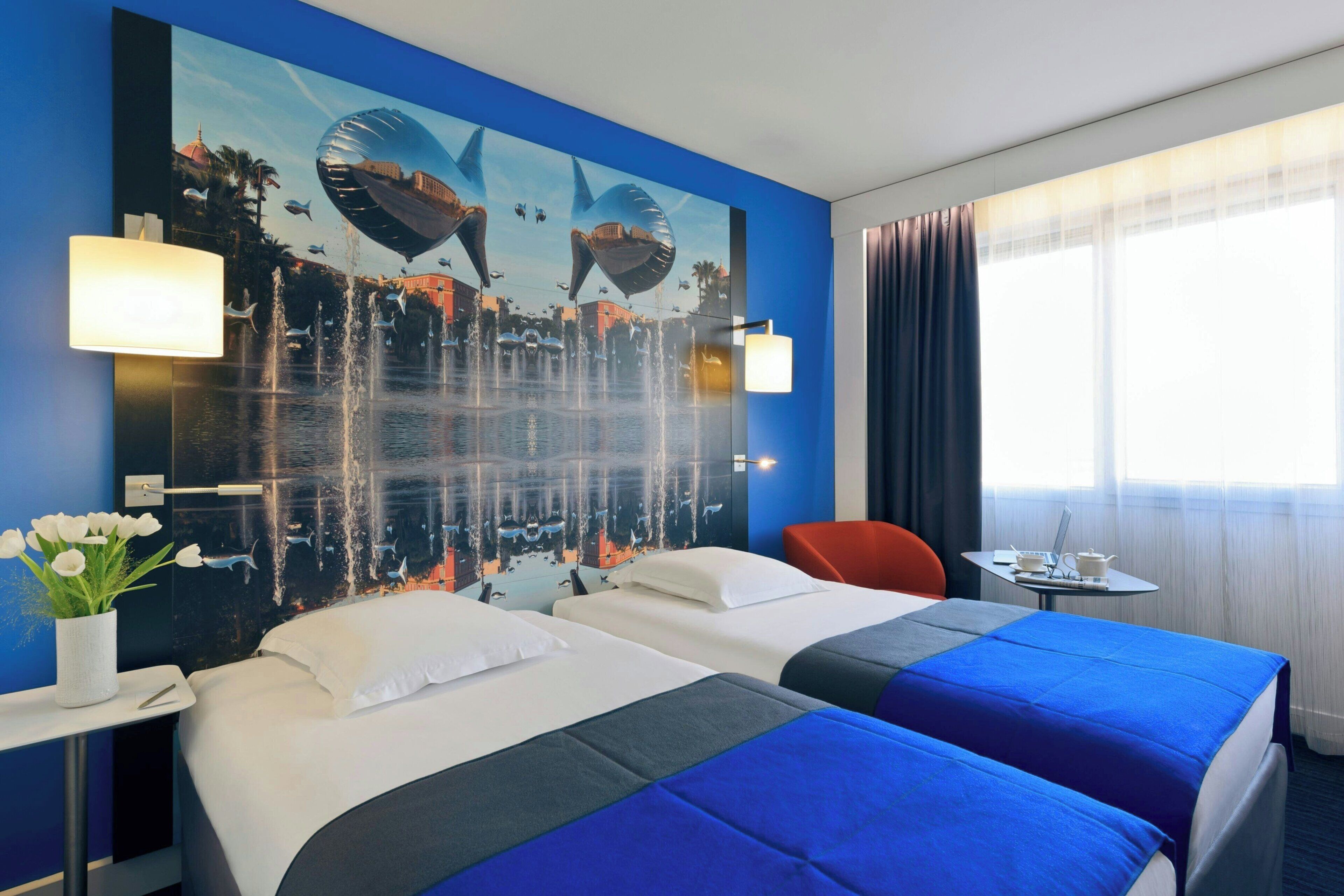 A hotel room with two beds, a large mural on the wall, a window with curtains, a small table with a laptop, and a vase of flowers.