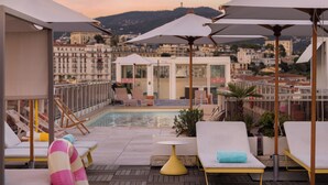 Zona de piscina en la azotea con sillones de descanso y sombrillas, con vistas a un paisaje urbano que incluye edificios y colinas.