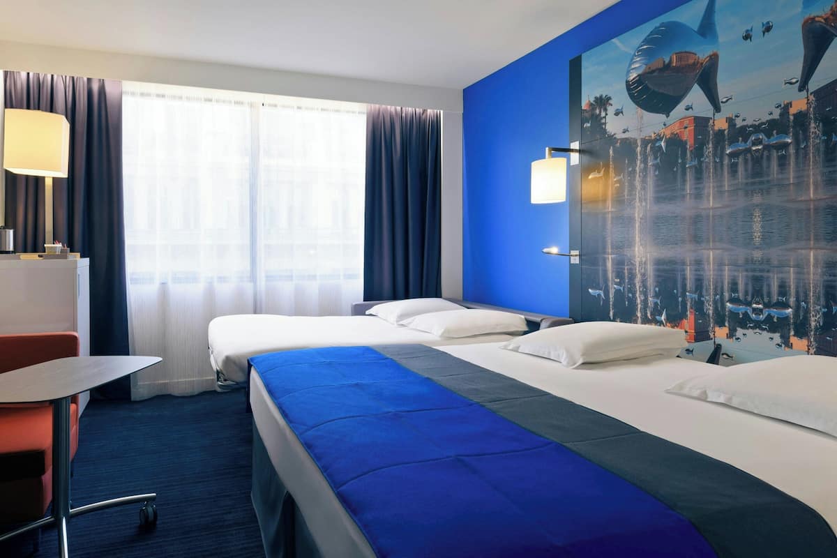 A hotel room with two beds, a desk, a chair, a large window with curtains, and a mural of sharks on the wall.