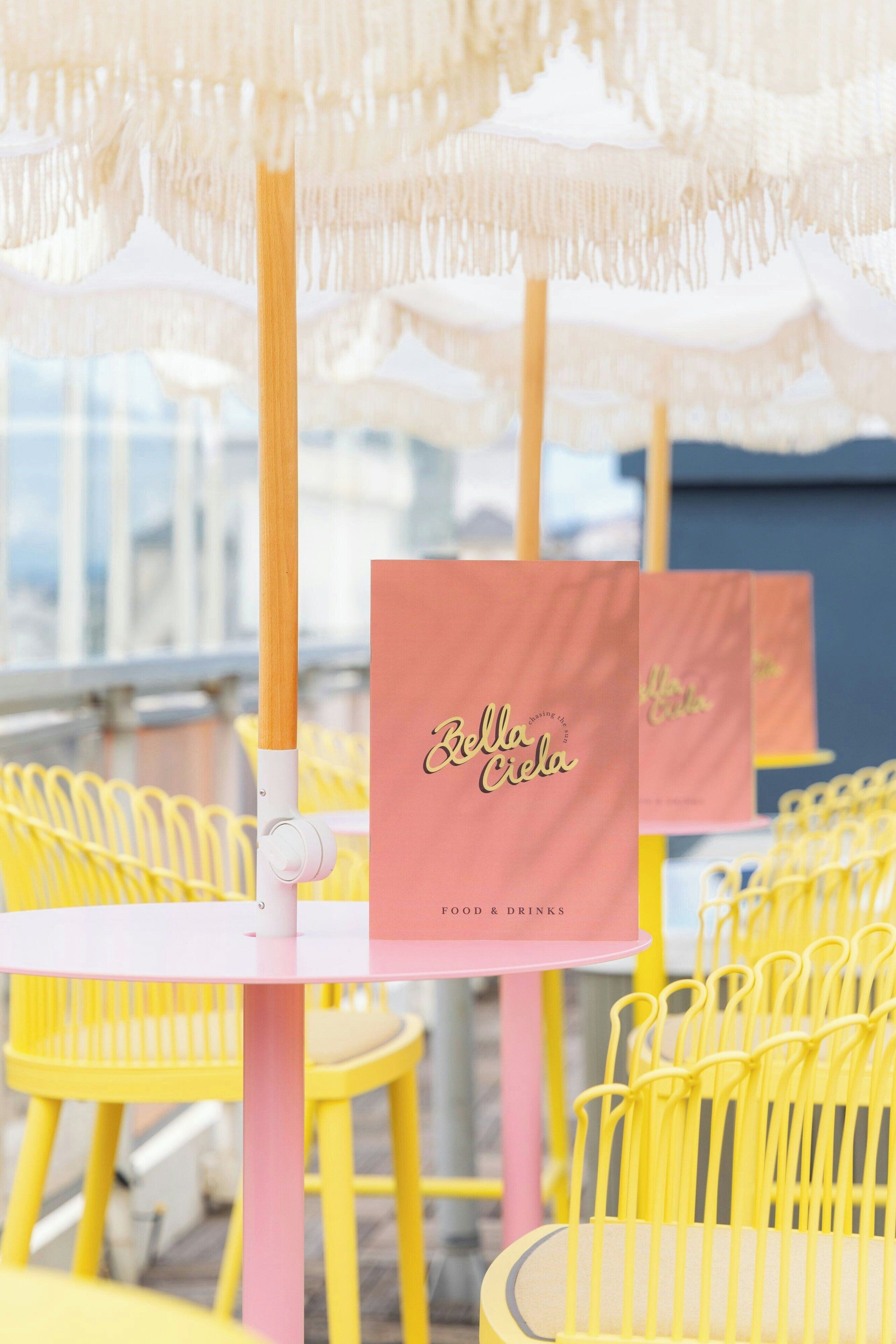 Outdoor seating with yellow chairs and tables, a white umbrella, and a pink menu stand with "Bella Crela" branding.