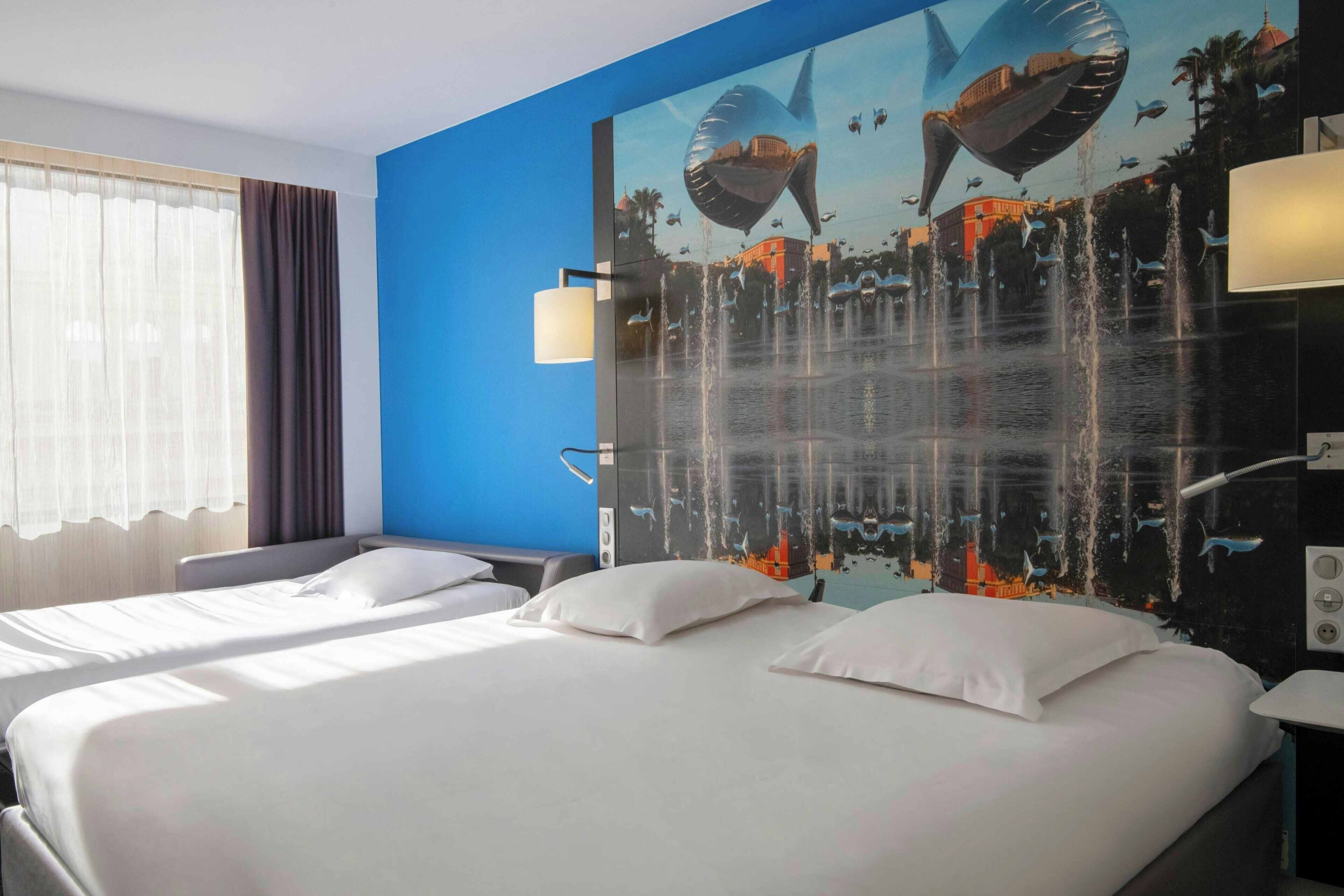 A hotel room with two beds, a large wall mural, a window with curtains, and a nightstand with a lamp.