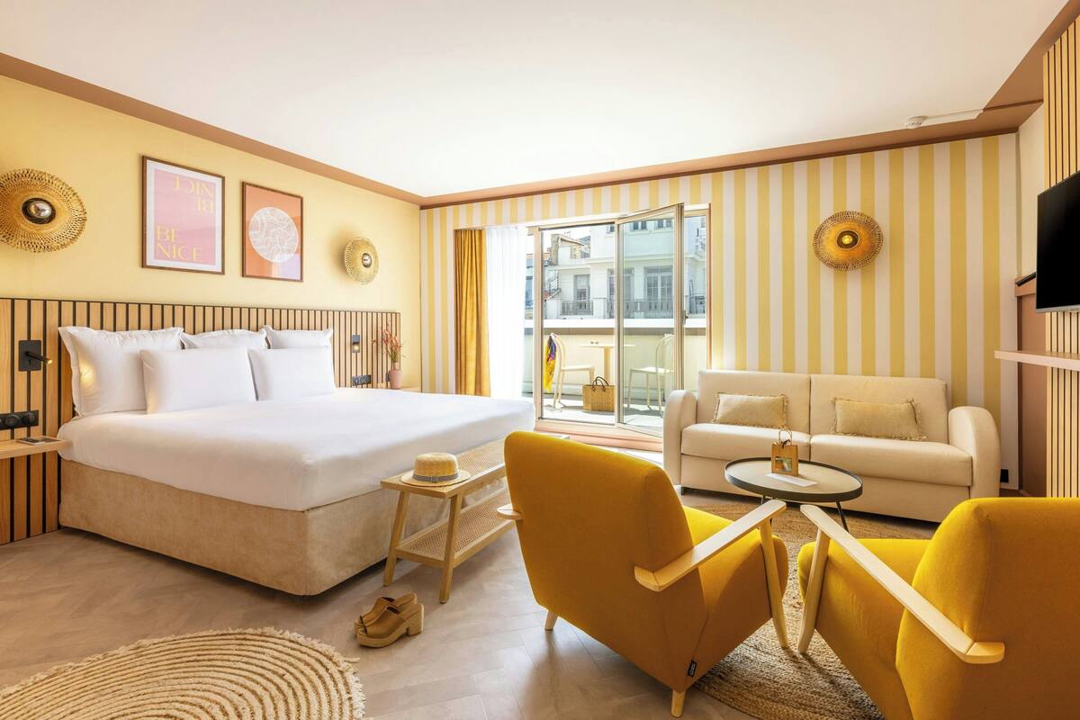 A hotel room with a large bed, a sofa, two yellow armchairs, a small table, and a TV.