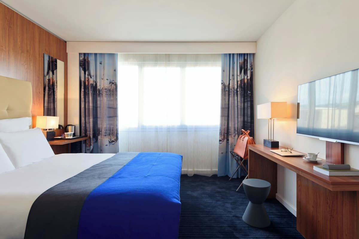 A hotel room with a large bed, a desk with a chair, a television, and a window with curtains.