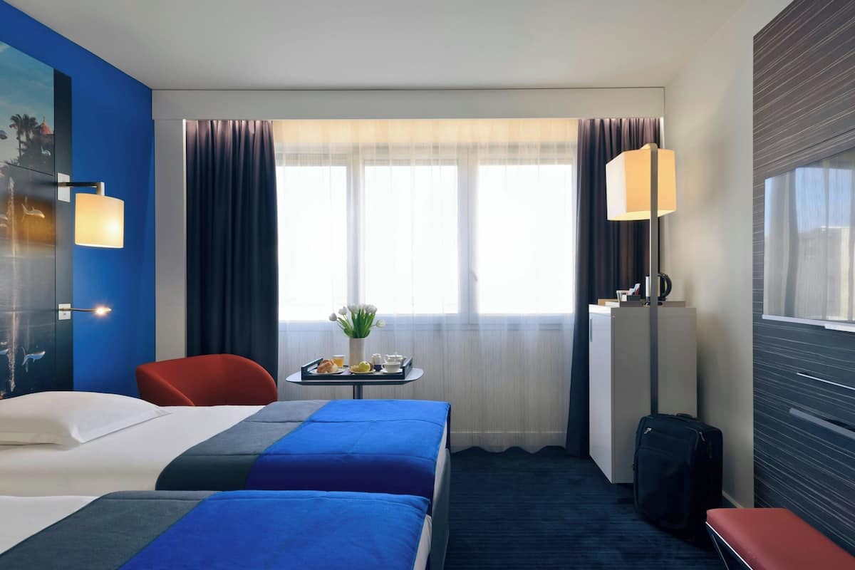 A hotel room with two beds, a desk with breakfast items, a chair, a suitcase, and a large window with curtains.