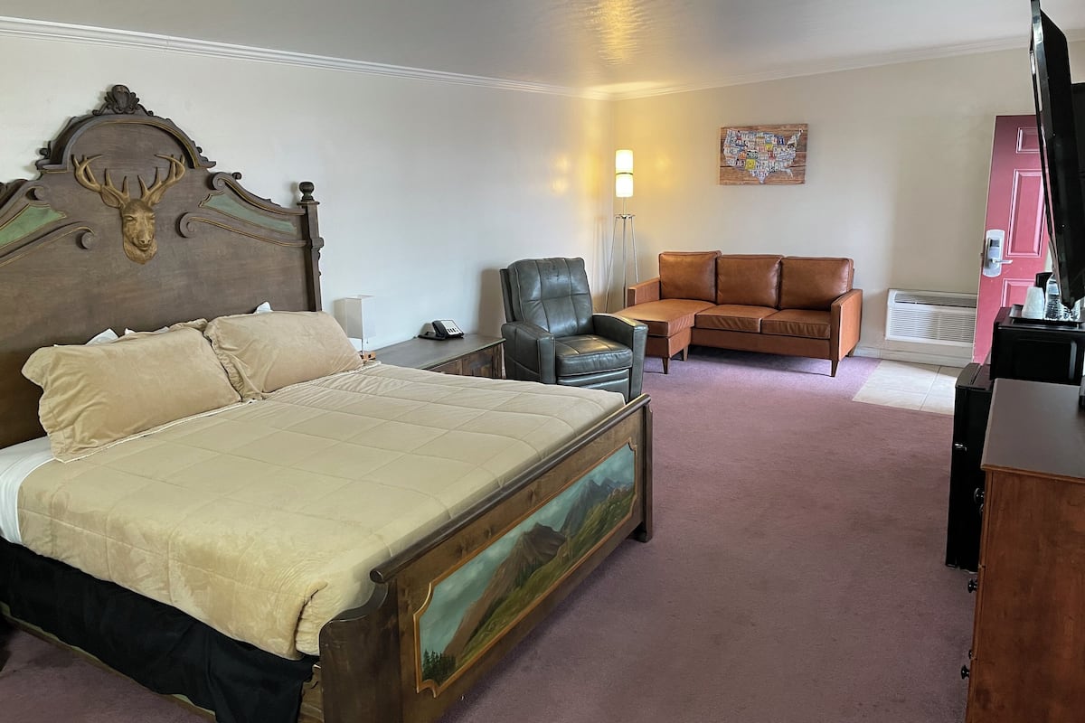 Une chambre d  ’hôtel avec un grand lit, une tête de lit en bois ornée d’une tête de cerf, un fauteuil en cuir, un canapé marron, un tableau accroché à la tête de lit, une lampe et une carte au mur.