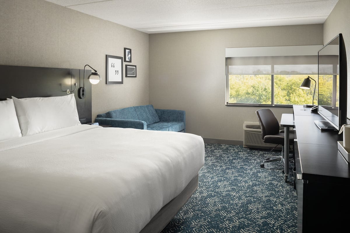 Hotelkamer met een groot bed, een bureau met computer, een stoel en een raam met uitzicht op bomen.