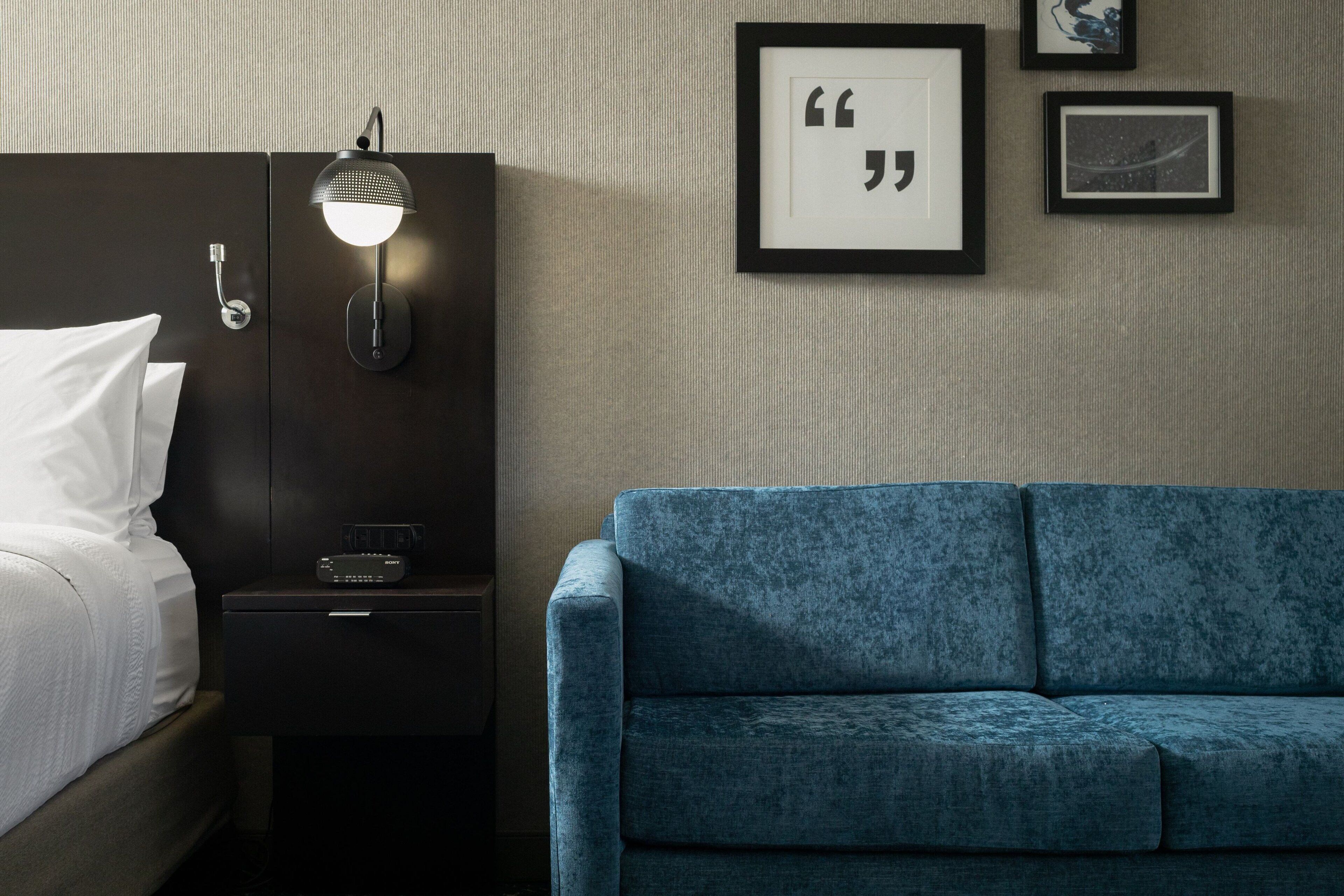 A hotel room with a bed, a nightstand with a lamp, a blue sofa, and framed quotes on the wall.