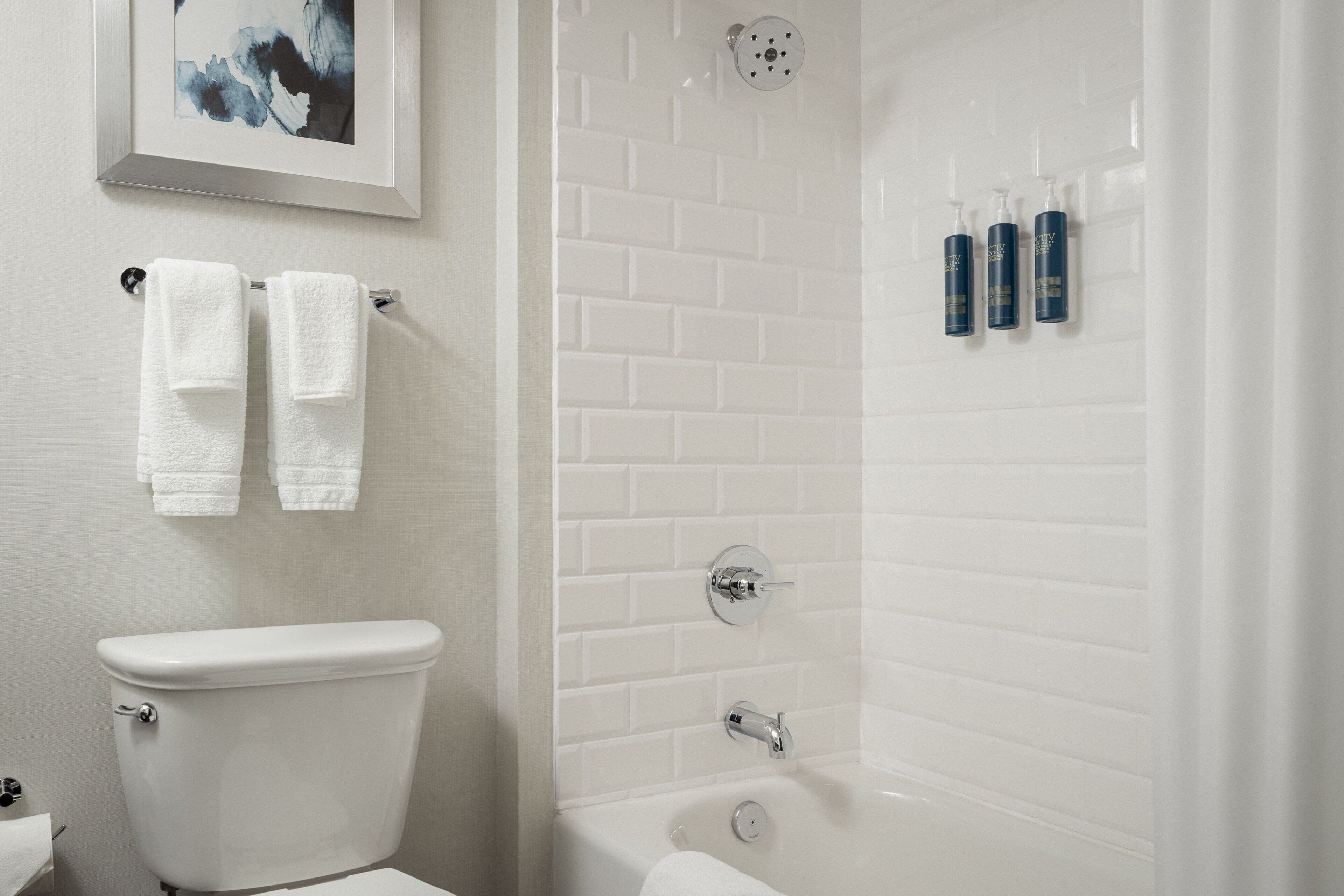 Un baño con un inodoro blanco, una ducha con pared de azulejos blancos y tres botellas de champú en la pared.