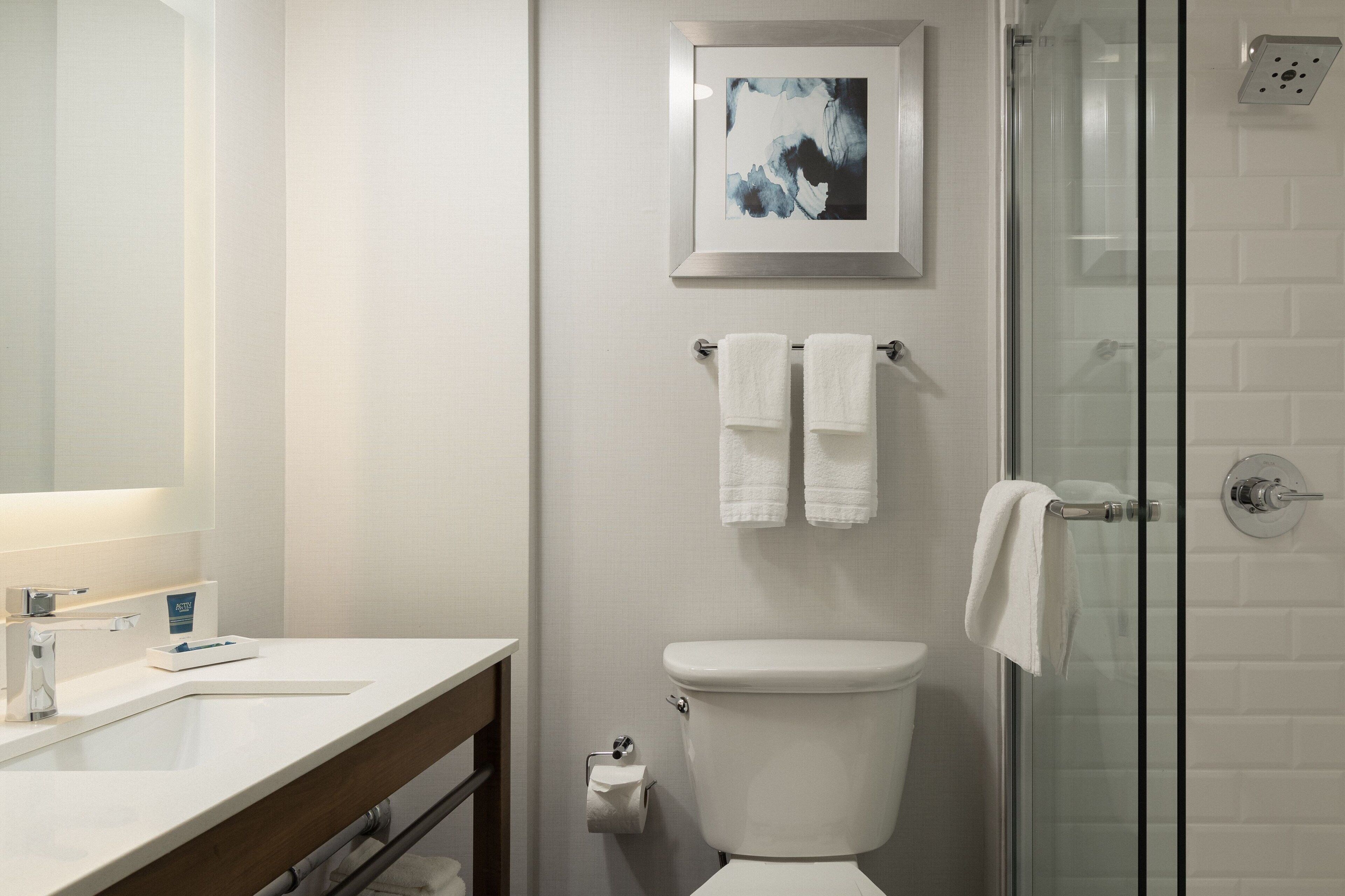 A modern bathroom with a white sink, a glass shower enclosure, and a framed abstract painting above the toilet.