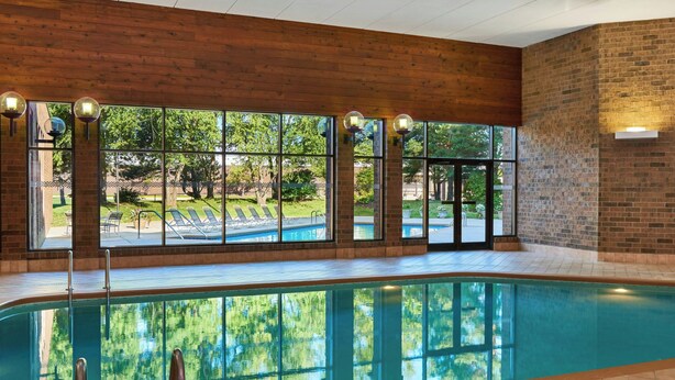 An indoor swimming pool with clear glass walls, a wooden ceiling, and a view of a green outdoor area. - Four Points by Sheraton Milwaukee North Shore (Milwaukee)