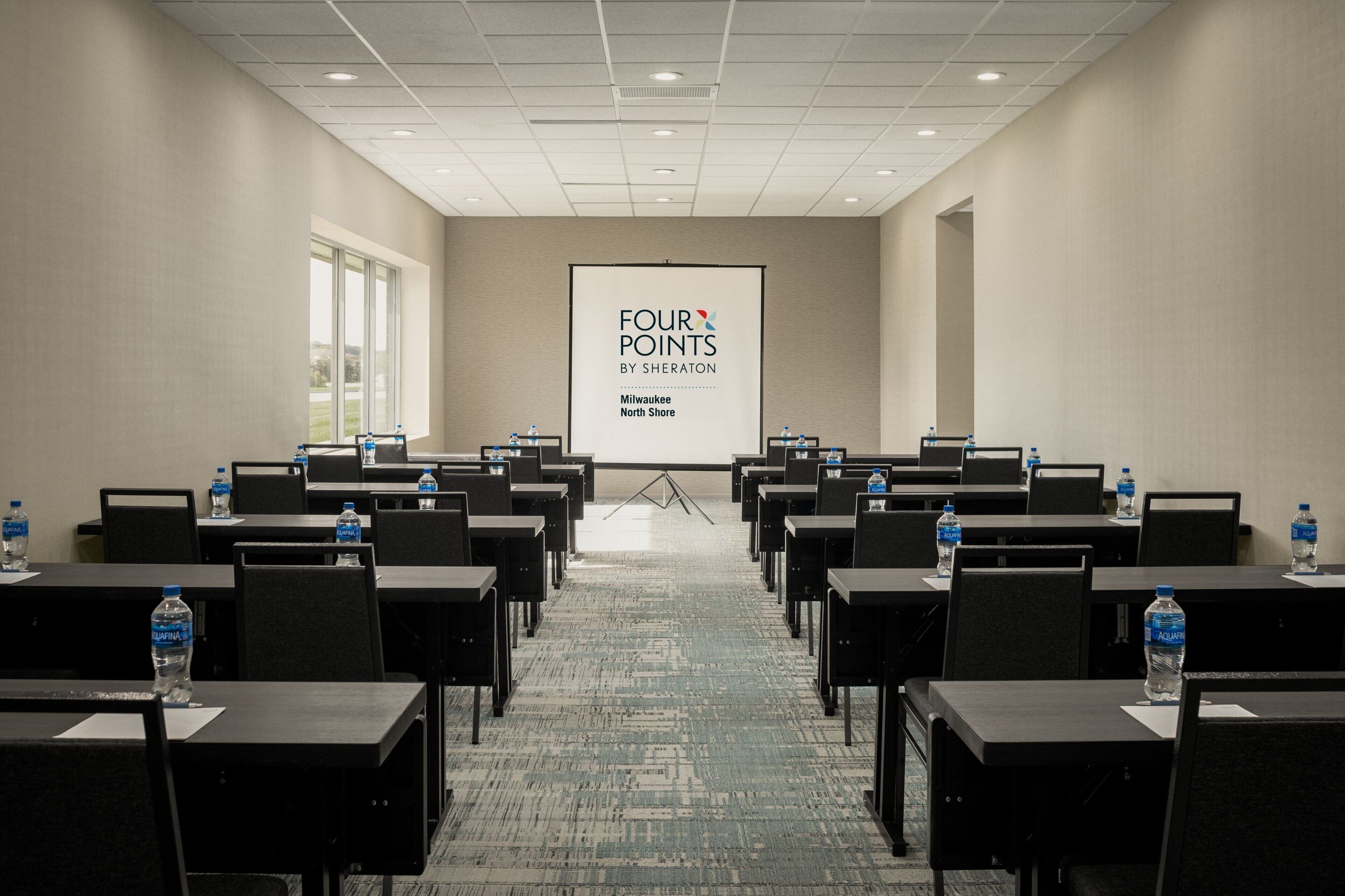 A conference room with rows of tables and chairs, a projector screen displaying "FOUR POINTS BY SHERATON", and water bottles on each table.
