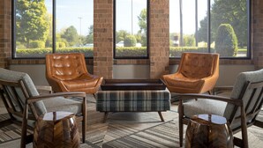 A seating area with leather chairs and a plaid ottoman, a brick fireplace, and large windows with a view of greenery and a parking lot. - Four Points by Sheraton Milwaukee North Shore (Milwaukee)