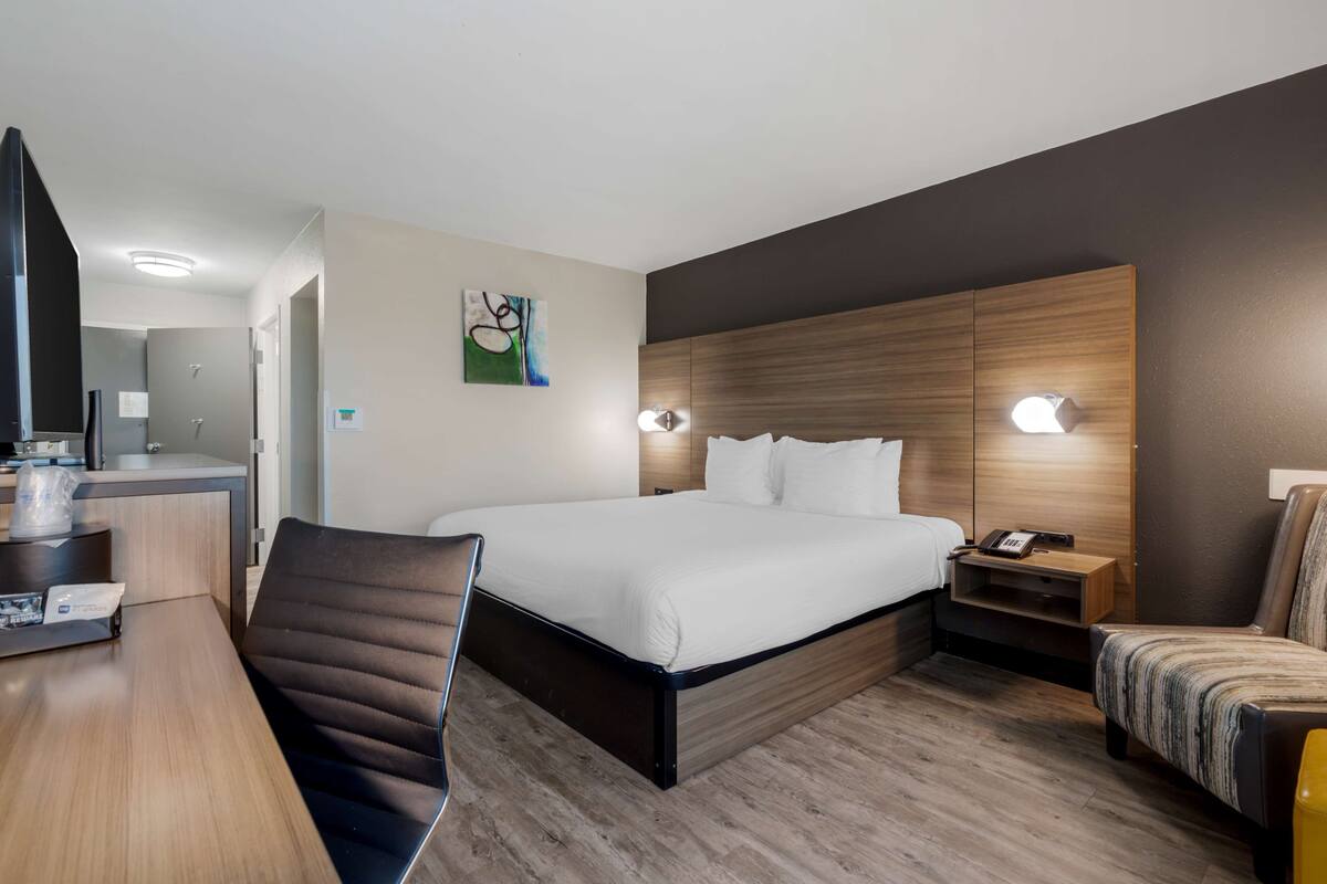 Quarto de hotel com uma cama grande, uma escrivaninha, uma cadeira, uma mesa de cabeceira e uma televisão fixada na parede.