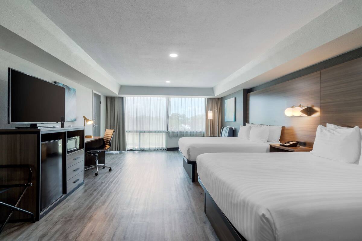 Quarto de hotel com duas camas, uma mesa de trabalho e uma televisão, além de uma janela grande com cortinas.