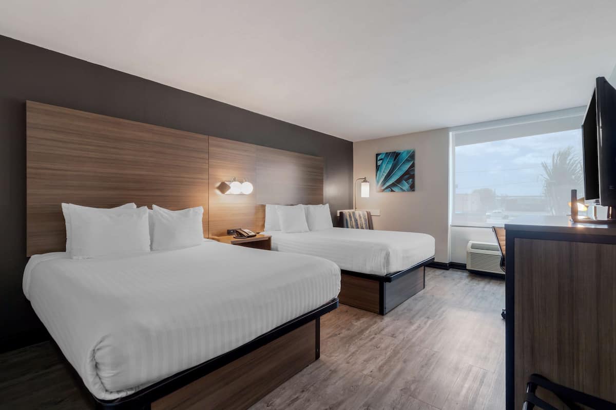 Quarto de hotel com duas camas, uma escrivaninha, televisão e uma janela grande.