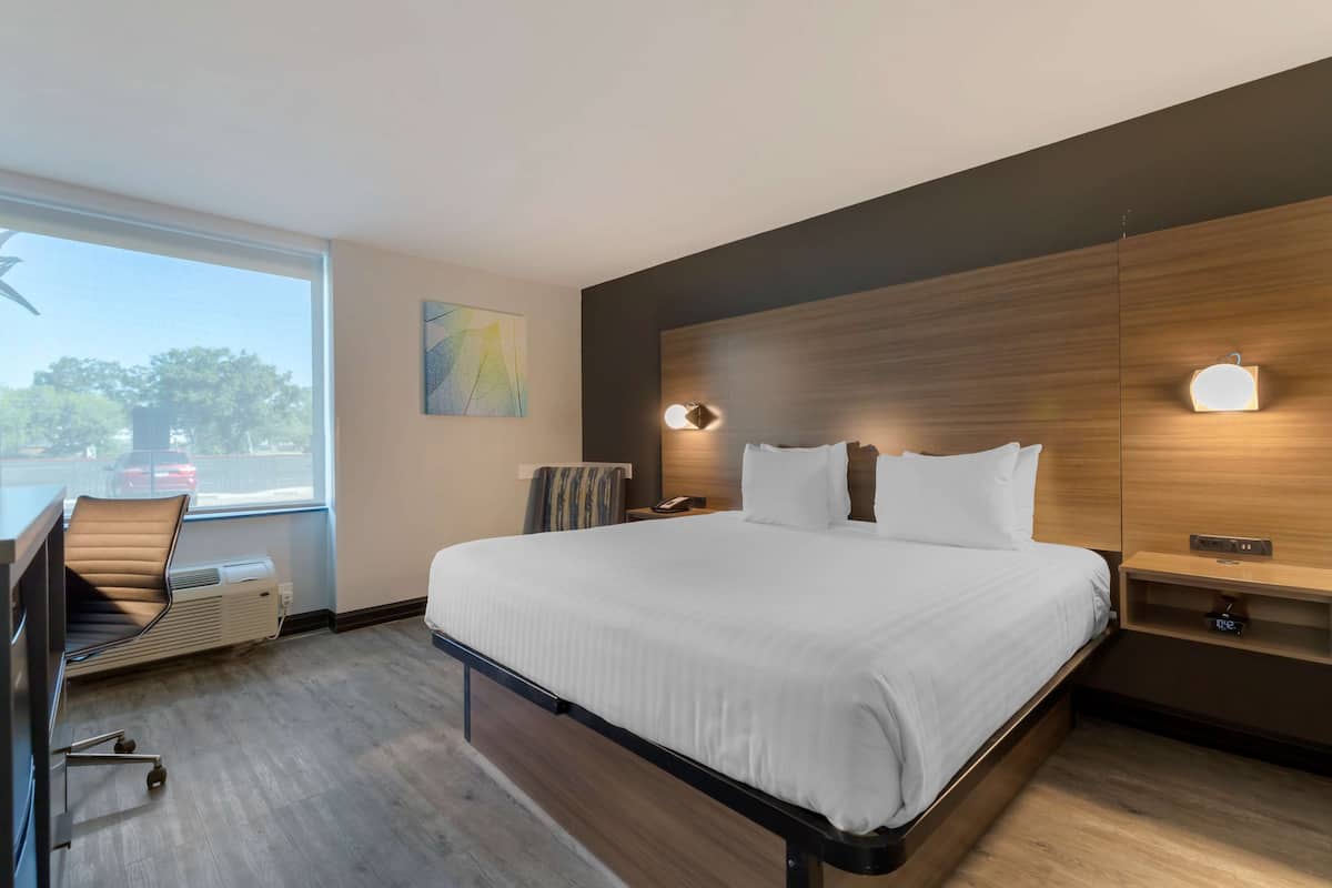 Quarto de hotel moderno com uma cama grande, uma escrivaninha com cadeira, uma janela com vista exterior e um candeeiro fixado na parede.