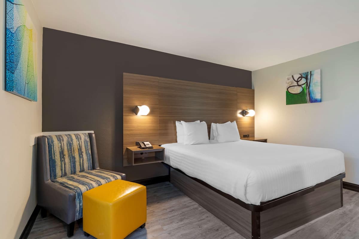 Quarto de hotel moderno com uma cama grande, mesa de cabeceira com telefone, uma cadeira e um pequeno banquinho.
