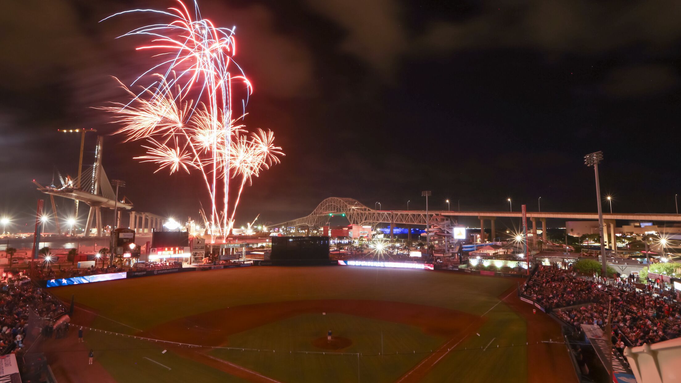 A baseball stadium with a large crowd, a bridge, and fireworks in the sky.
