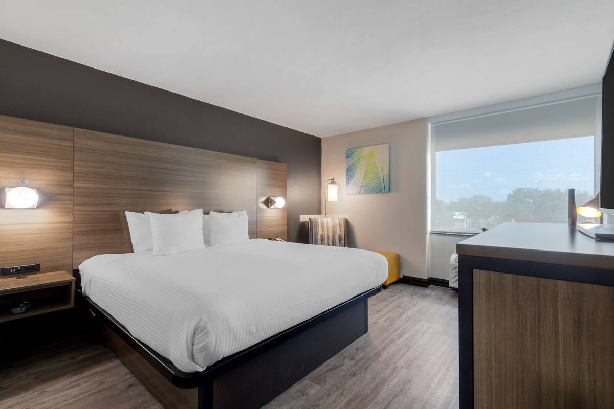 Quarto de hotel moderno com uma cama grande, abajures, uma pequena escrivaninha e uma janela ampla com vista para a cidade.