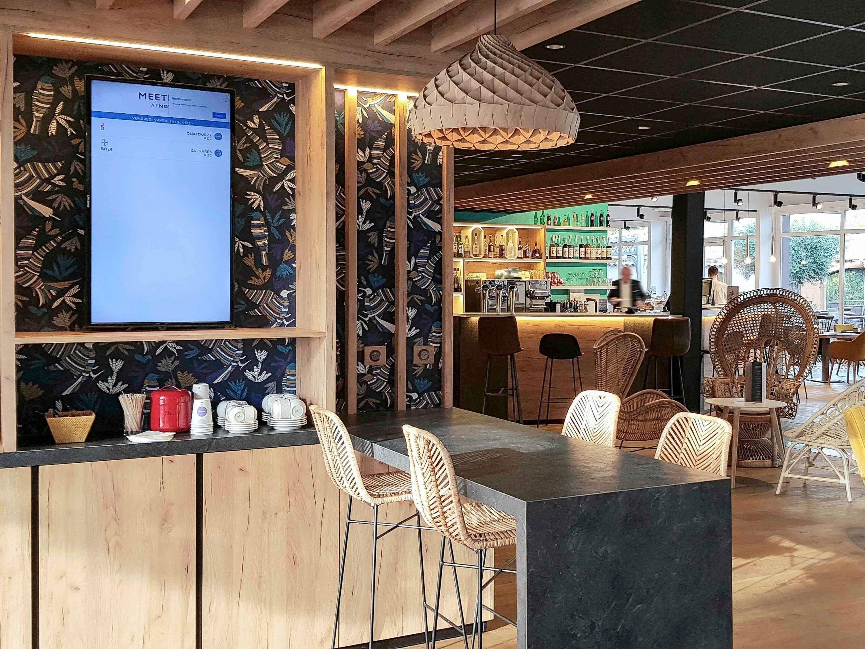 Un espace bar avec un comptoir en bois, des chaises en osier et un grand écran affichant le menu.