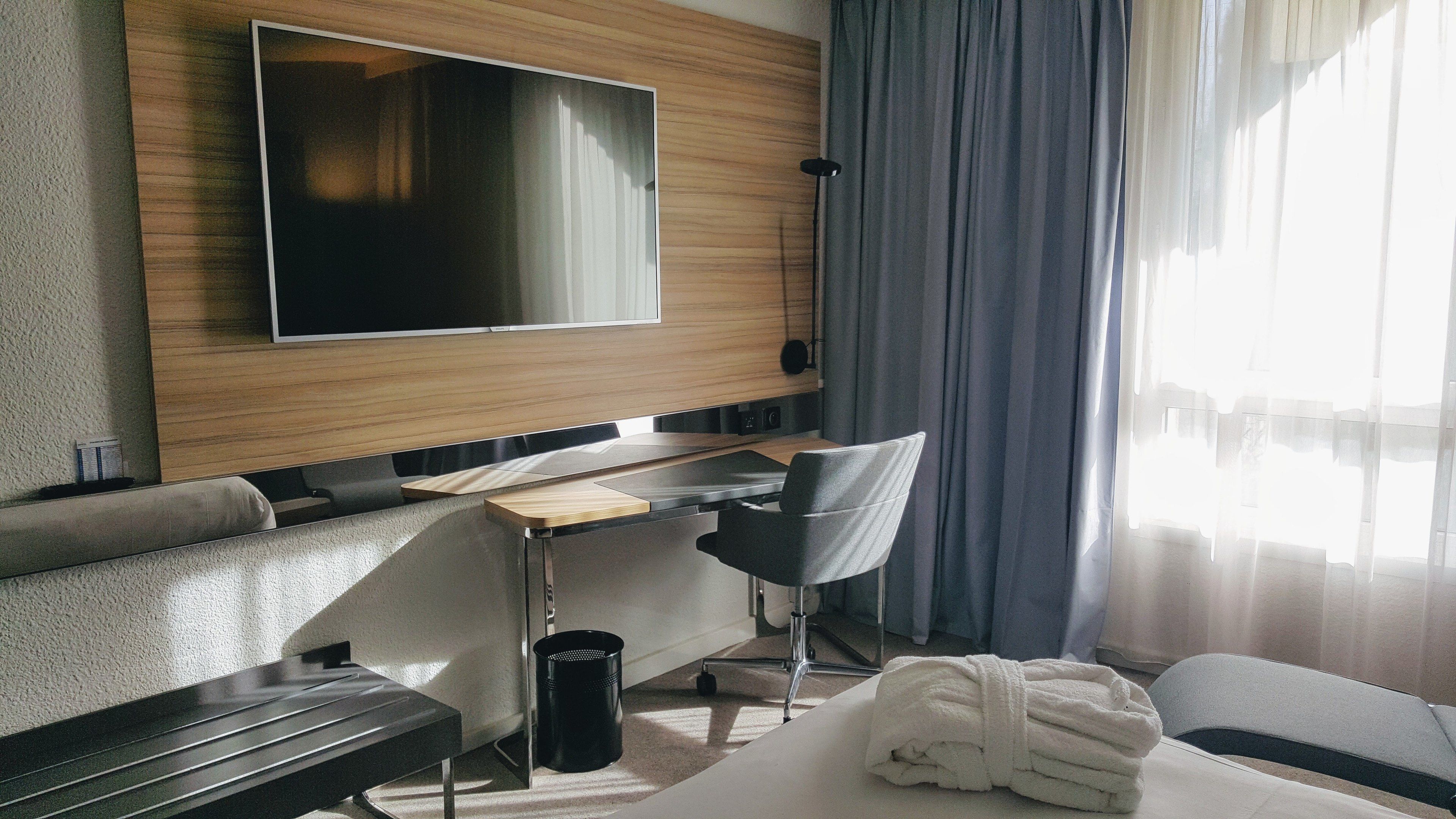 A modern hotel room with a flat-screen TV mounted on the wall, a desk with a chair, a sofa, and a large window with curtains.