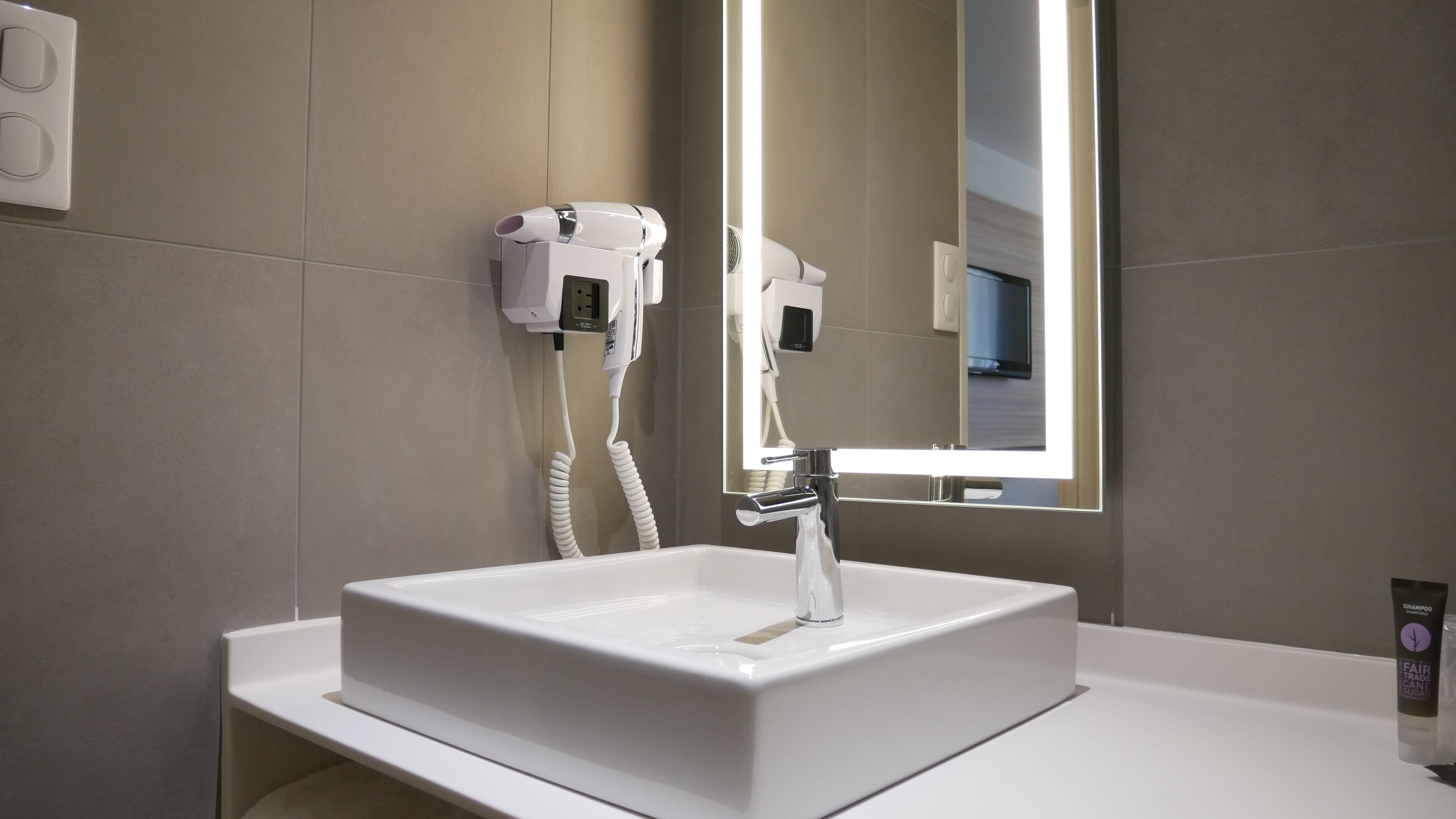 A modern bathroom with a white sink, a hairdryer, and a mirror with integrated lighting.