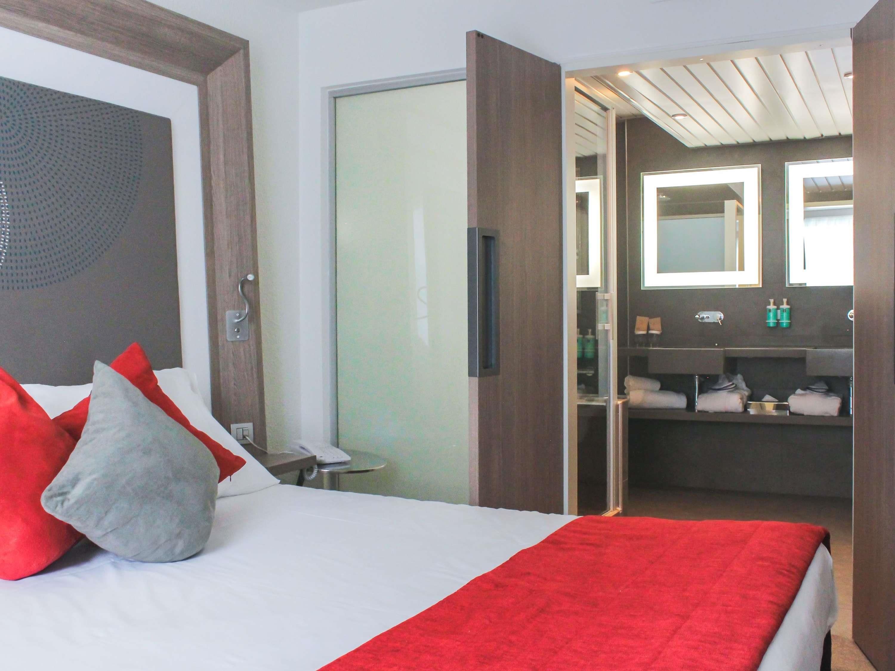 Une chambre d’hôtel avec un lit, des oreillers, une couverture rouge, une salle de bain avec un miroir et un lavabo.