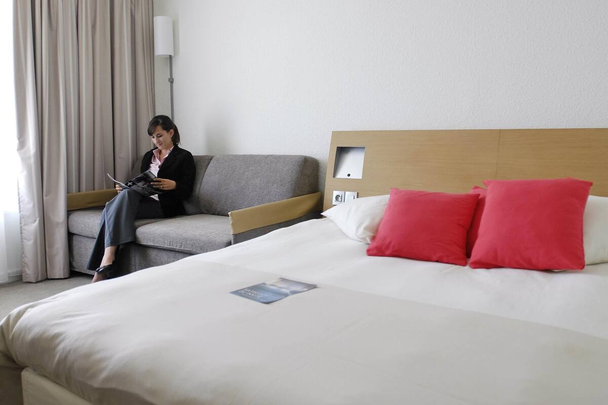A hotel room with a bed, a sofa, a lamp, and a person reading a magazine.