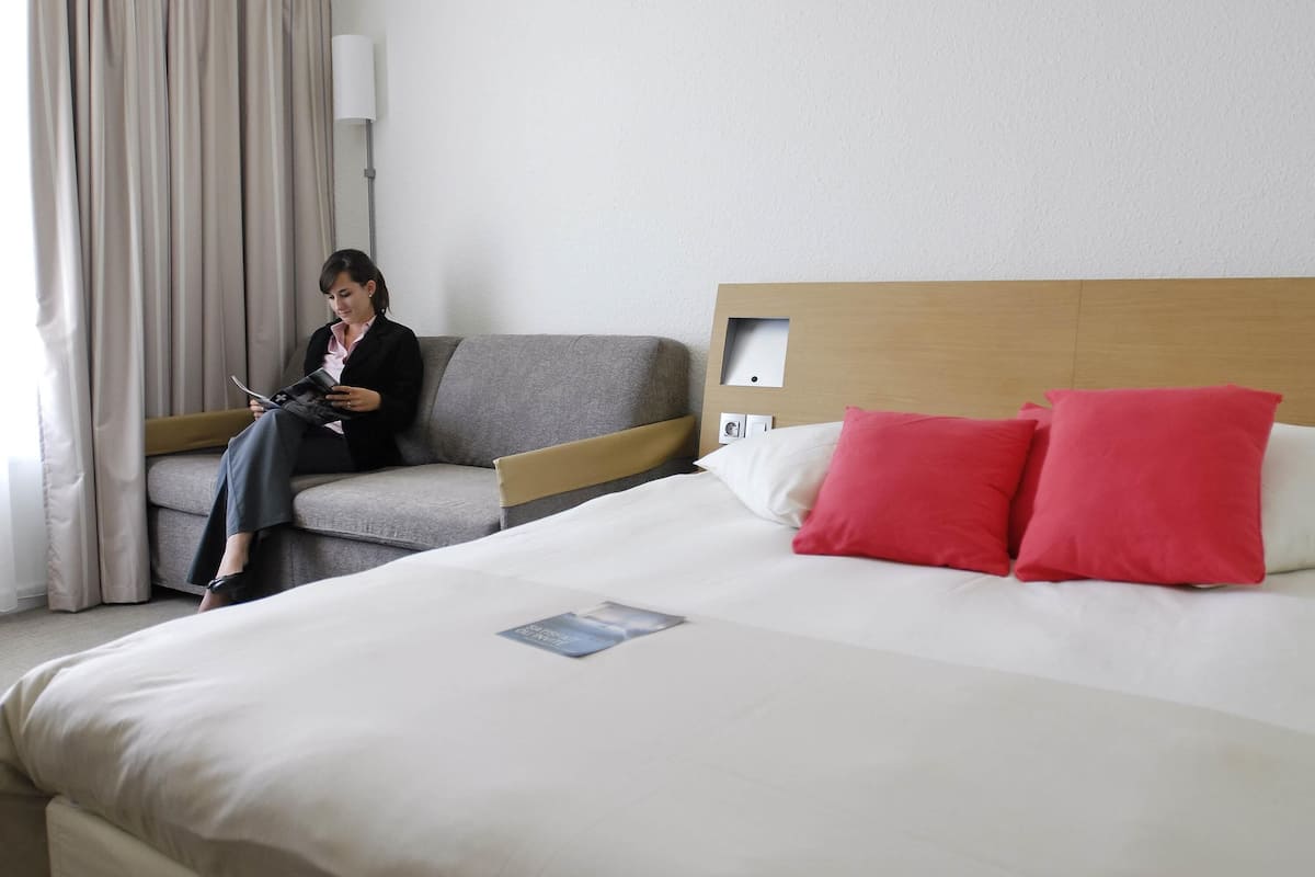 Ein Hotelzimmer mit einem Bett, einer Couch, einer Lampe und einer Person, die eine Zeitschrift liest.