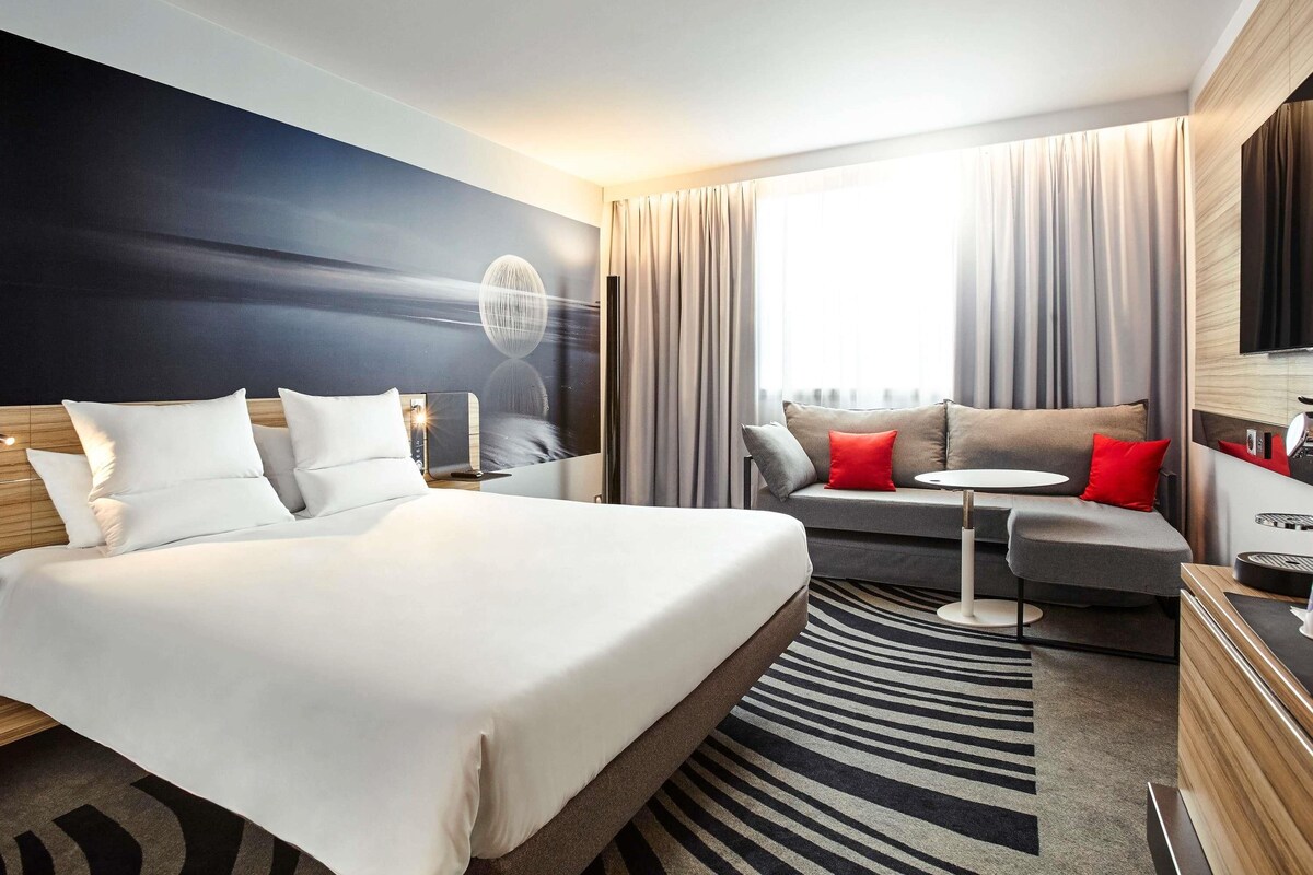 A modern hotel room with a large bed, a sofa, a small table, and a TV.