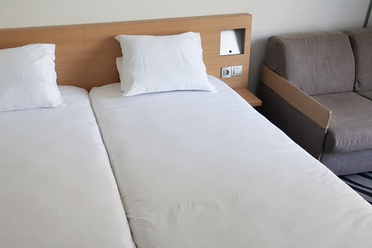 Una cama doble con sábanas blancas, un cabecero de madera, un teléfono en la mesita de noche y un sillón marrón.
